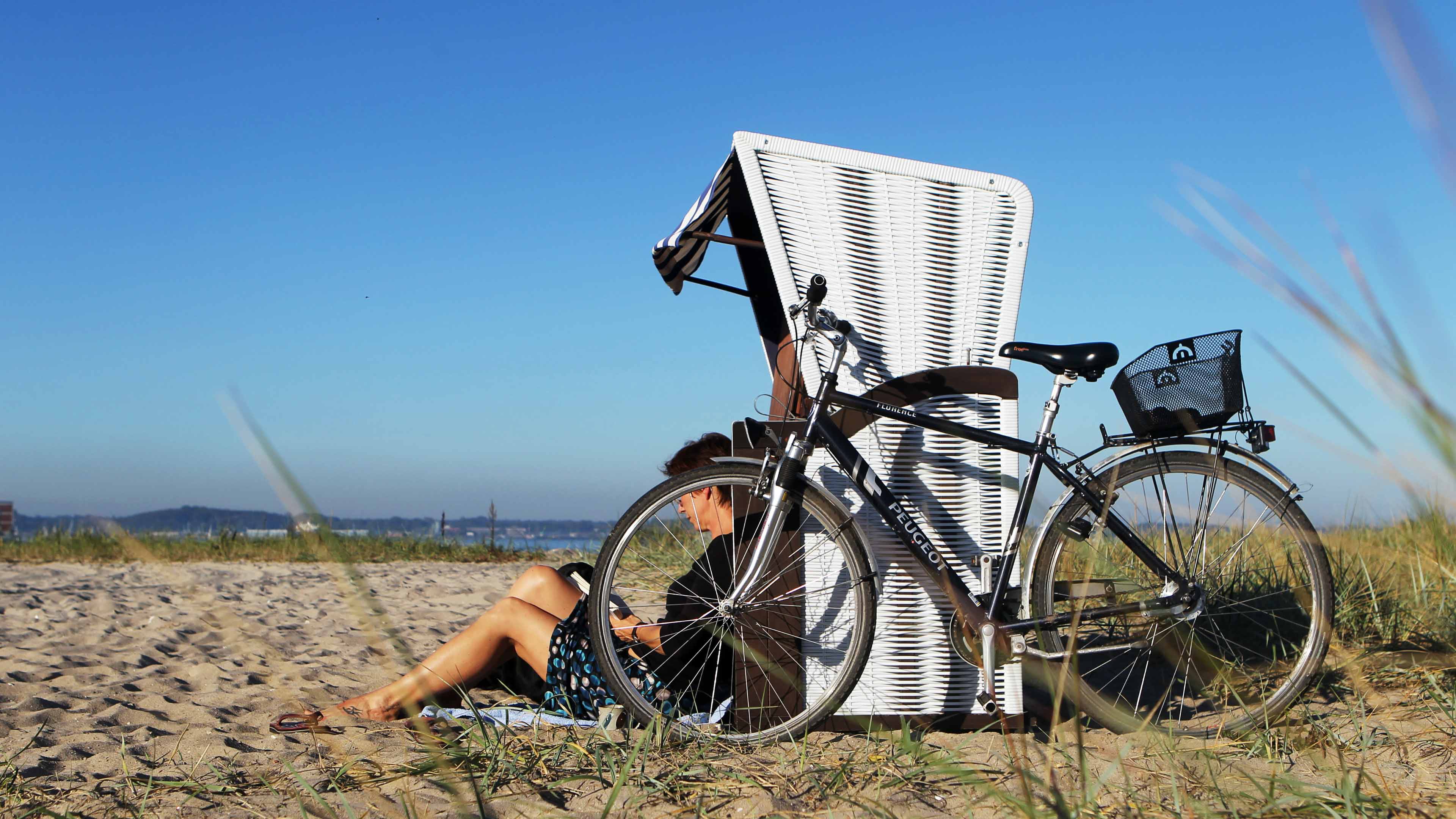 Fahrrad lehnt an weißem Strandkorb, davor sitzt eine lesende Frau