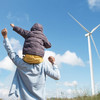 Blick von hinten auf eine Person mit Kind auf den Schultern vor einer Windkraftanlage