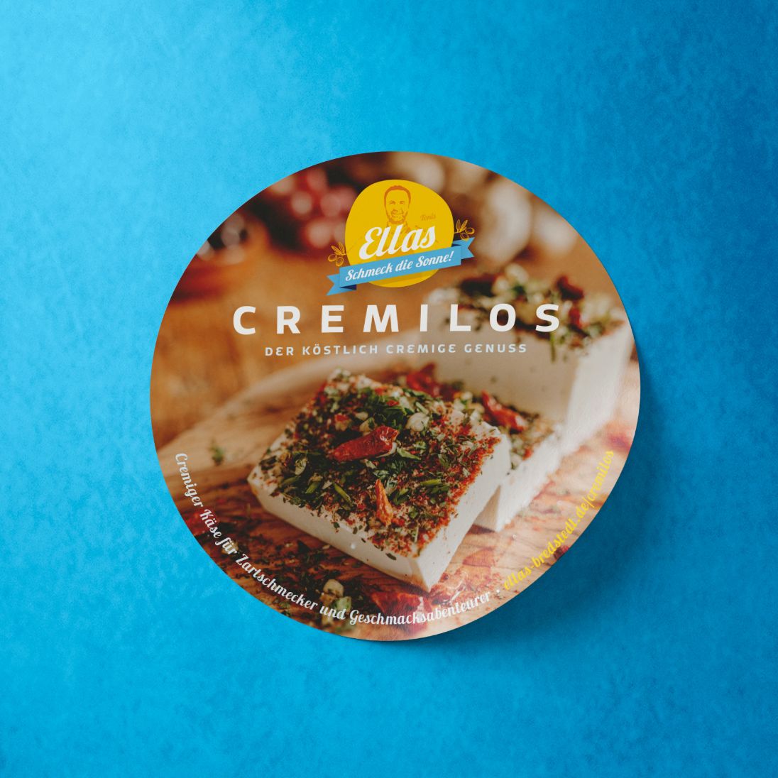 h2_ellas_teaser_03.jpg Ein runter Cremilos Aufkleber mit einem Weichkäse mit Kräutern und Chilis auf blauem Hintergrund