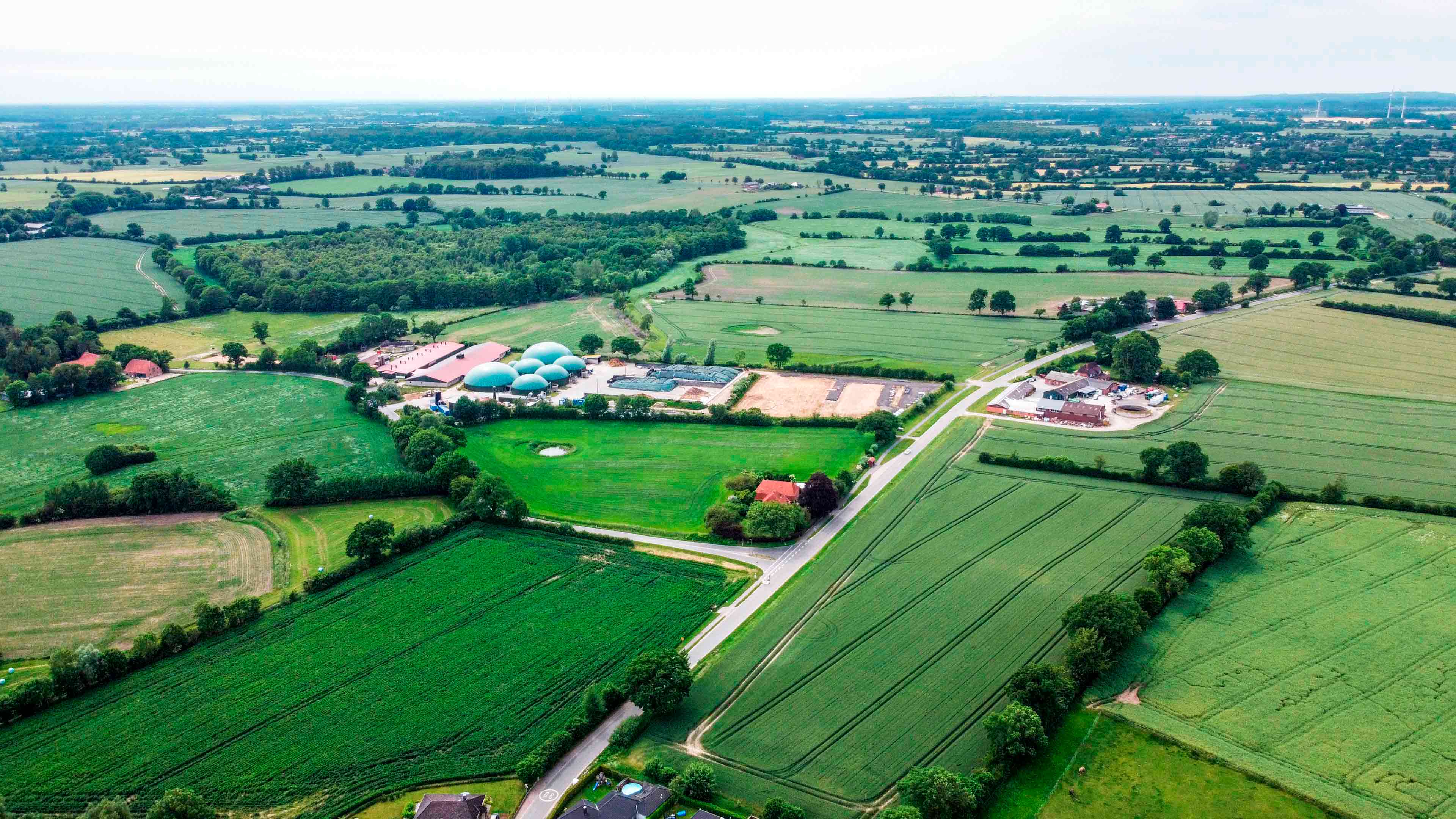 Luftaufnahme zeigt Biogasanlage, Wiesen und Bäume und eine Landstraße