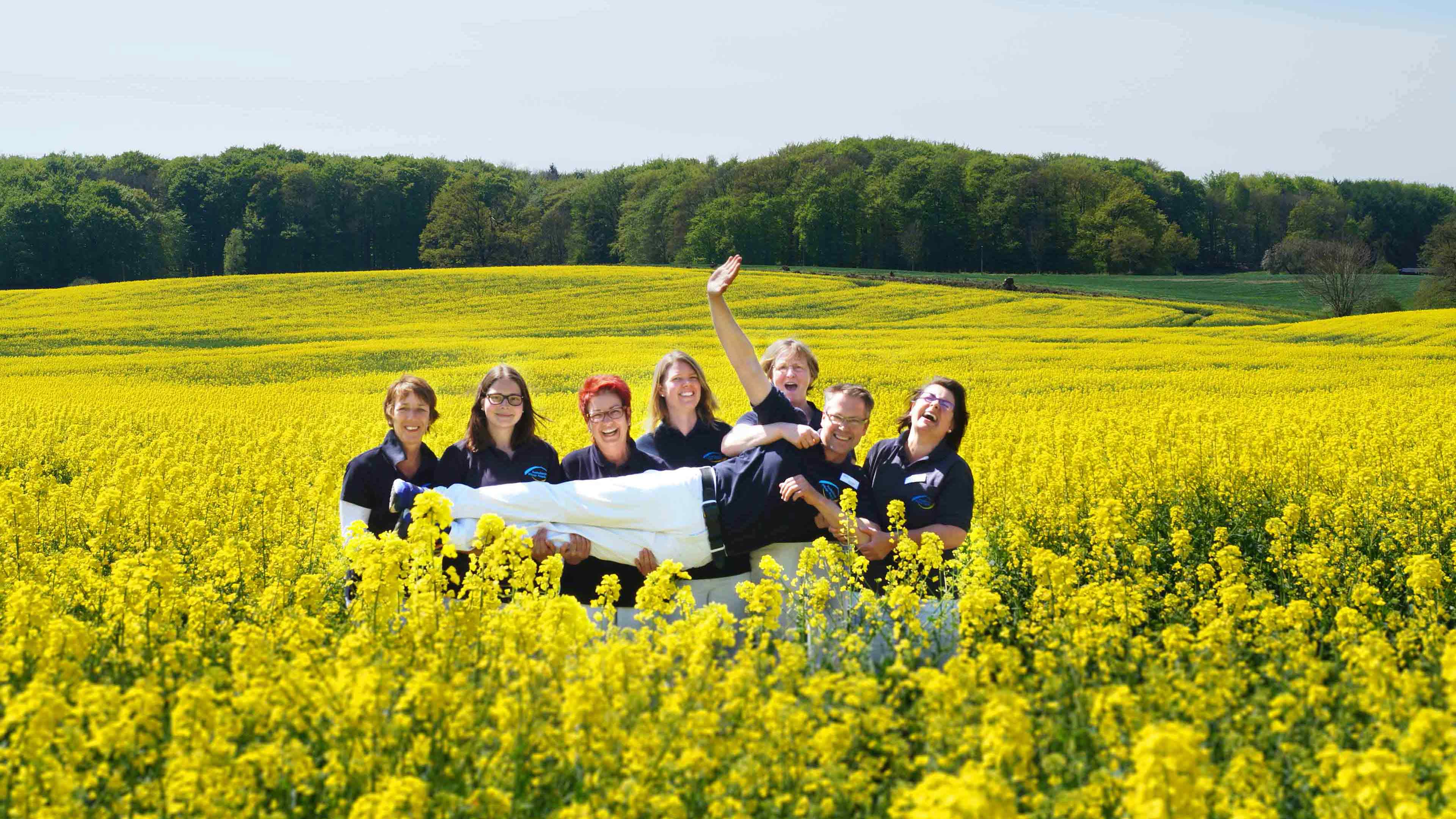 Sechs lachende Mitarbeiter stehen im gelben Rapsfeld und halten eine Person quer vor sich hoch