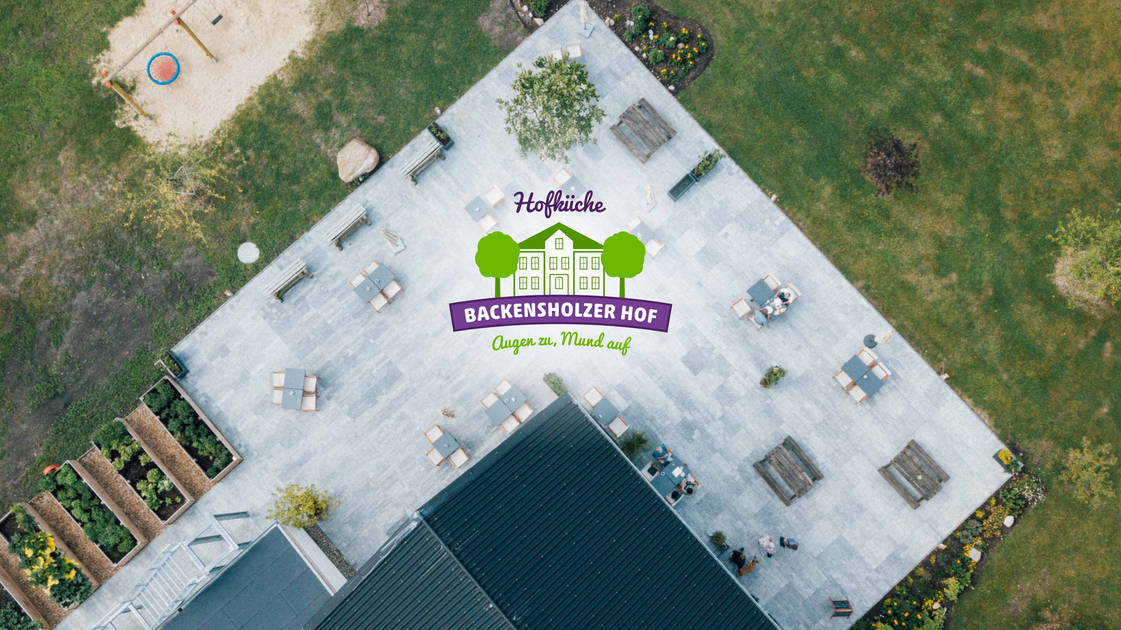 Draufblick auf Terrasse, Hochbeete und Spielplatz rund um die Hofküche mit Logo des Backensholzer Hofs