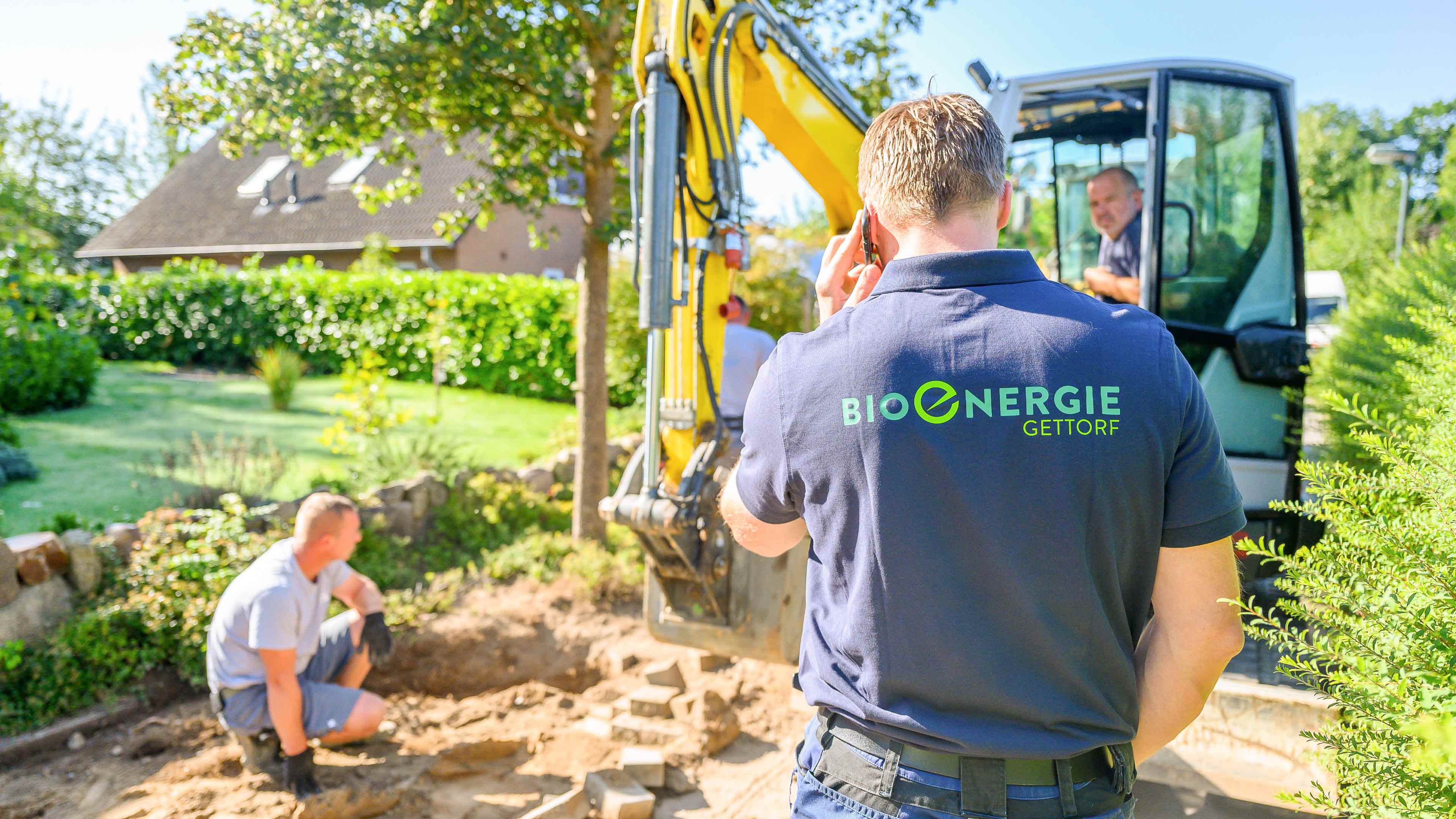 Rechteckiges Bild einer Person mit blauem Poloshirt mit Bioenergie Gettorf Logo steht vor einem kleinen Bagger und telefoniert