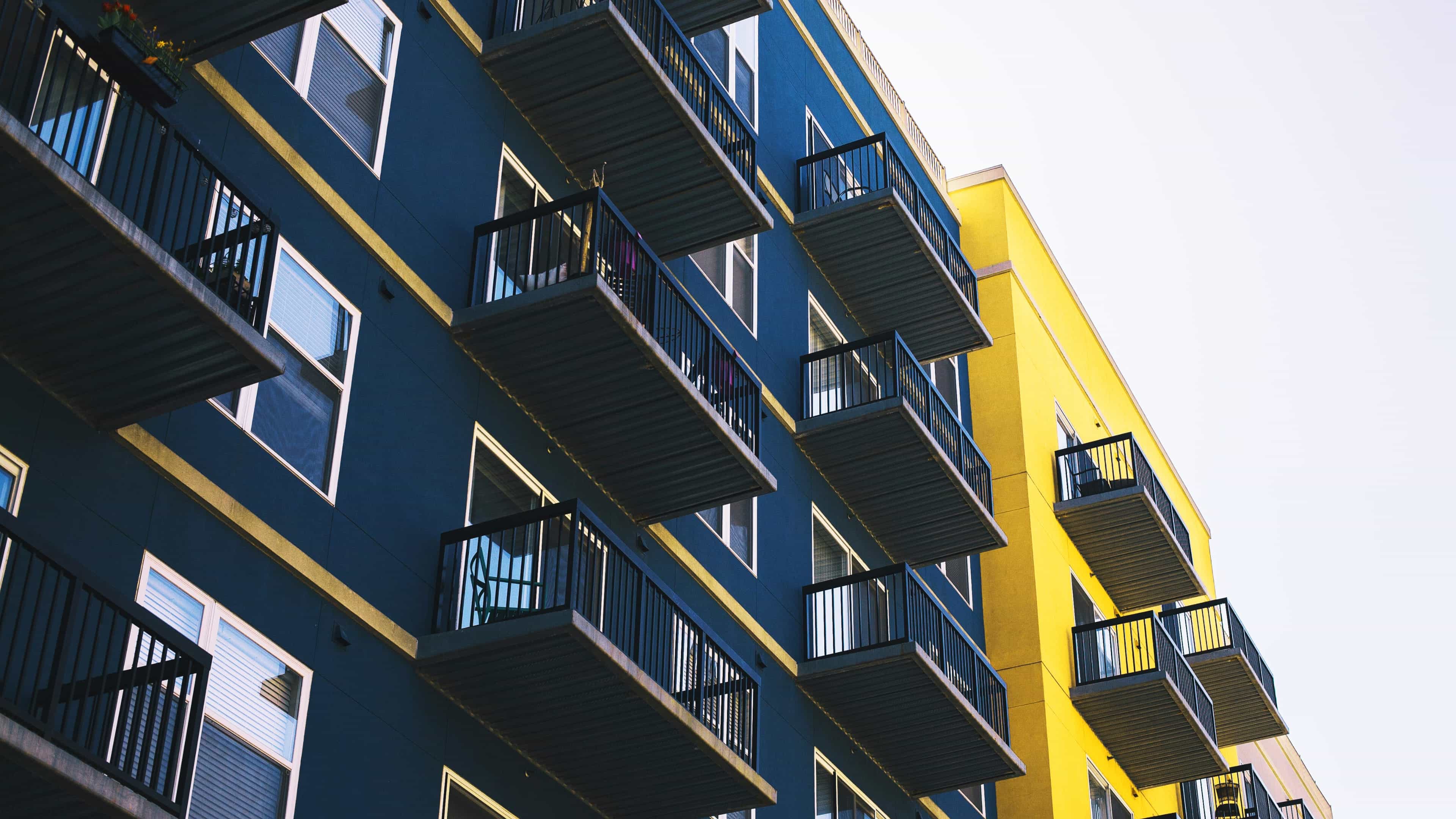 Blick hinauf auf ein blau-gelbes Wohnhaus mit vielen Etagen und Balkonen