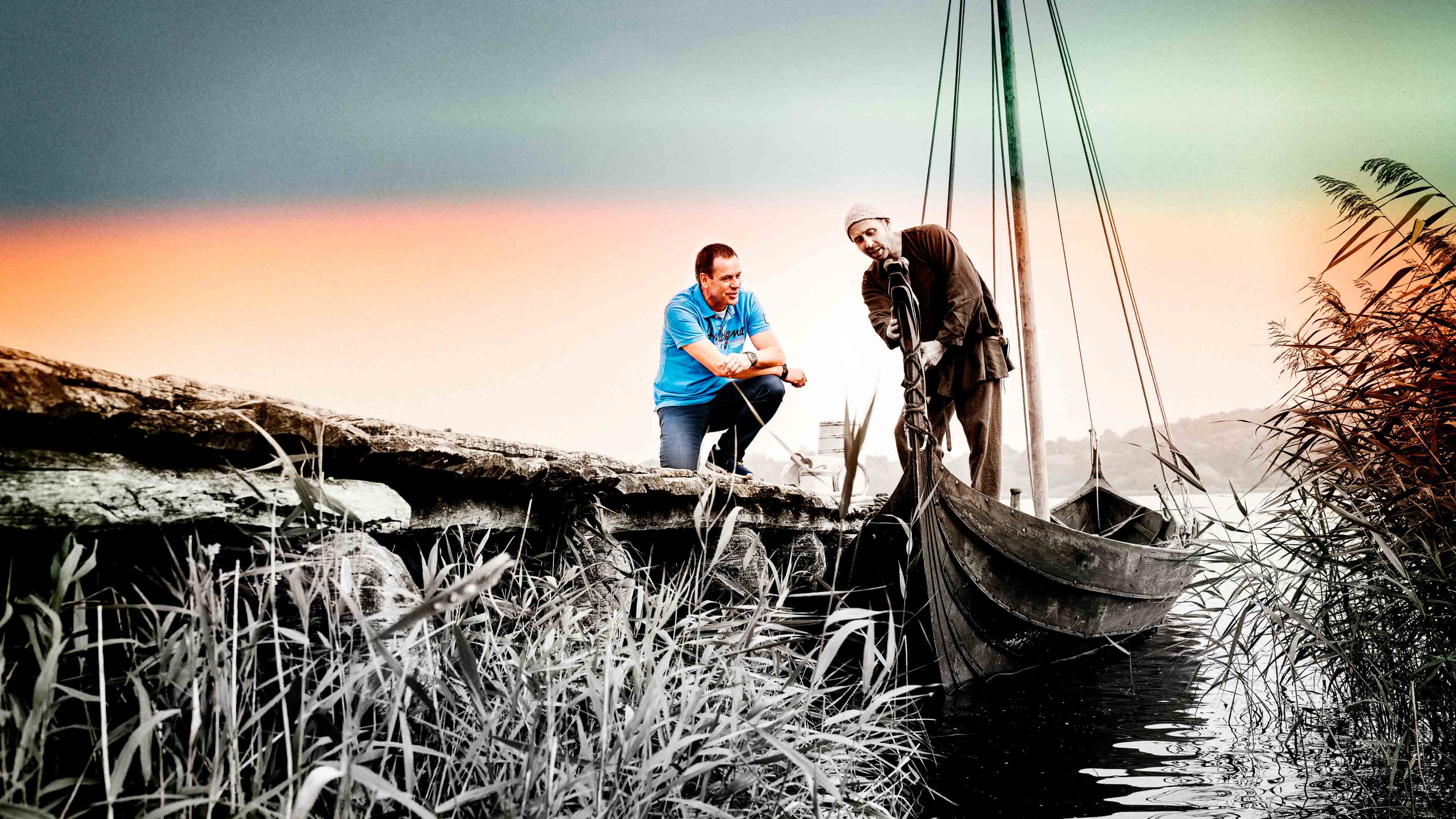 Moderner Mann hockt auf Steg und ein Mann zeigt ihm als Wikinger verkleidet ein Holzboot 