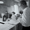 Schwarz-weißes Foto zeigt zwei am PC sitzende Mitarbeiter und einen über einem geöffneten Aktenordner stehend 