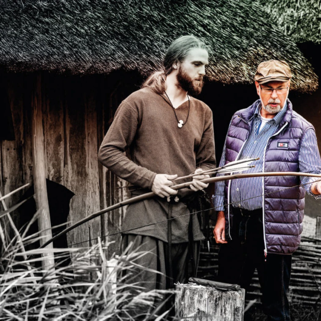h2_grenzuebergreifendes-marketing_teaser_10.jpg Bearbeitetes Foto eines Mannes, der draußen vor einer historischen Hütte in Haithabu steht und etwas erklärt. Neben ihm eine schemenhafte Darstellung eines Wikingers.