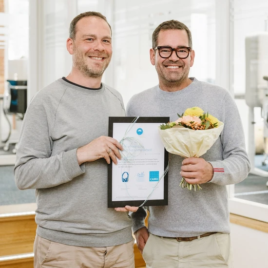 5 Jahre kreatives Vergnügen Foto von Jan-Henrik mit Chef Felix. Beide tragen einen hellgrauen Pullover und eine beige Hose. Jan-Henrik hält eine Urkunde, Felix einen Blumenstrauß.