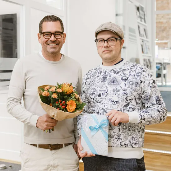 Zwei HOCHZWEI Mitarbeiter lächeln in die Kamera. Der eine hält einen Blumenstrauß in der Hand, der andere ein Geschenk.