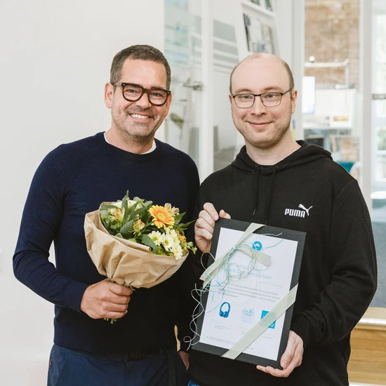 5 Jahre Code, Köpfchen & Schnack Foto von Tim und Chef Felix. Beide tragen einen dunklen Pullover. Tim hält eine Urkunde, Felix einen Blumenstrauß.