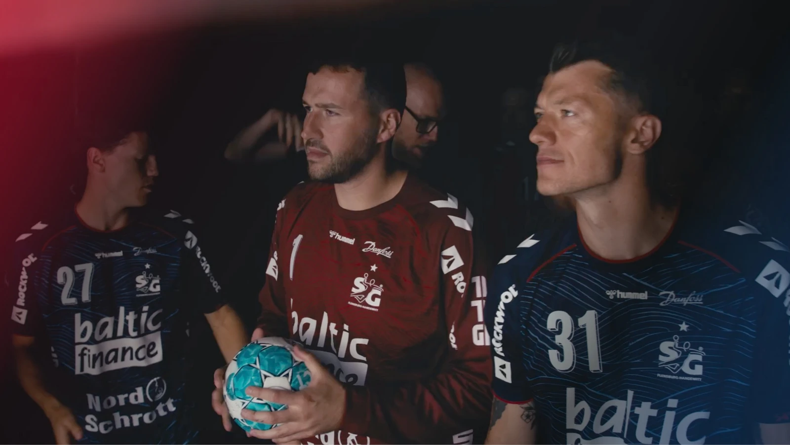 h2_sg_einlauftrailer_teaser_06.jpg Foto der SG Spieler im Halbdunklen. Vorne Burić mit dem Ball in der Hand und Jakobsen neben ihm