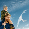 Bärtiger Mann mit Kleinkind auf den Schultern vor blauem Himmel und Syltumriss aus Wolken