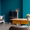 In einem blauen Zimmer steht ein Holztisch mit Blumenvase, Regal und ein Bett ist erkennbar