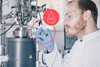 Ein Laborant steht vor seinen Arbeitsgeräten und hält ein Laborglas mit einer Flüssigkei in seiner Hand. Eine Visualisierung veranschaulicht, dass es sich um 