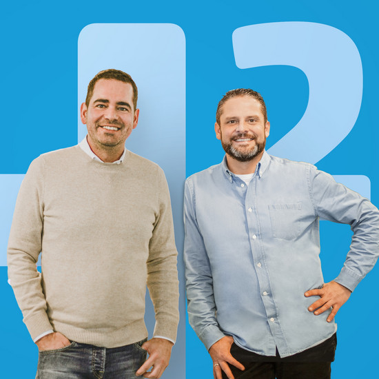 Das Geschäftsführerduo Felix Worm und Timo Klaas stehen lächelnd und mit entspannter Körperhaltung vor einer blauen Wand. Auf dieser ist ein H2-Aufdruck zu sehen. 