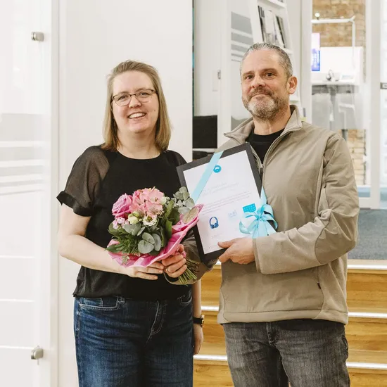 Zwei Personen lächeln in die Kamera, die Frau trägt einen Blumenstrauß in der Hand und der Mann eine Urkunde