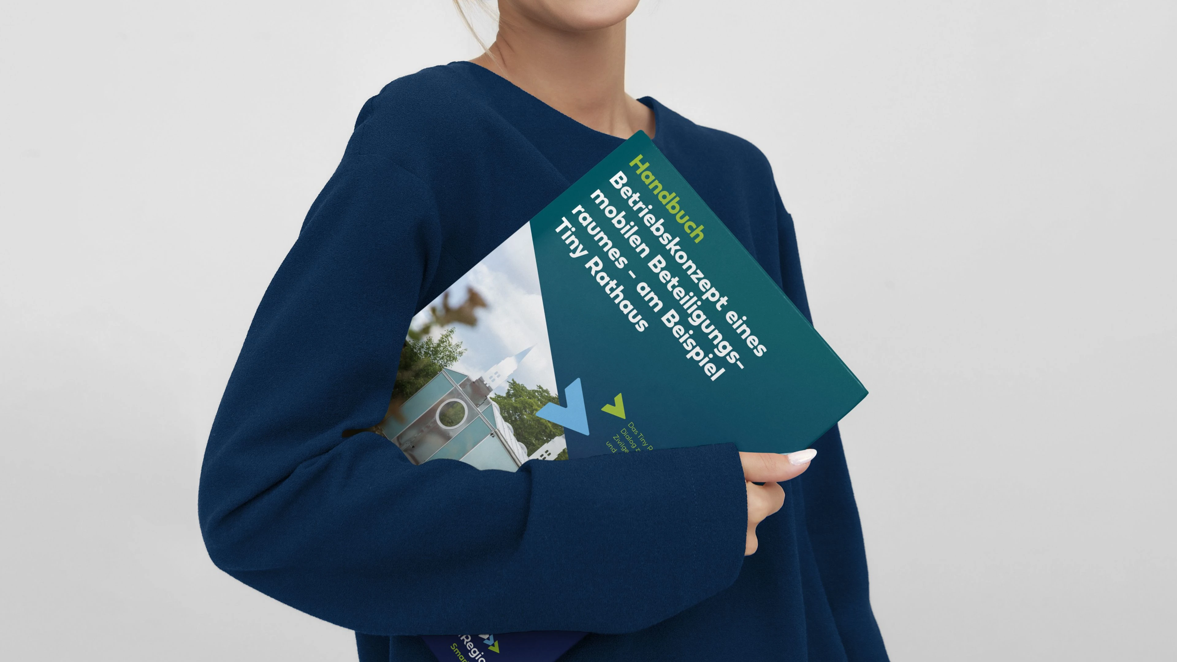 hochzwei_kielregion-handbuecher_slider_01.jpg Eine Person hält ein Handbuch mit dem Titel Betriebskonzept eines mobilen Beteiligungsraumes - am Beispiel Tiny Rathaus