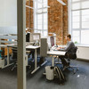 Blick in ein Büro mit drei Arbeitsplätzen, ein Mitarbeiter sitzt am Schreibtisch; hohe weiße Fenster und Klinkerwand